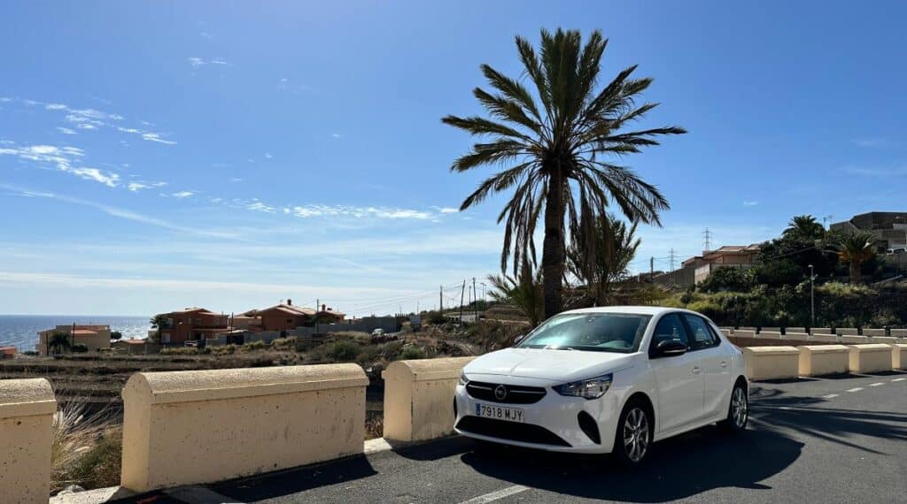  Weißer Opel Corsa als Mietwagen auf Küstenstraße im Süden Teneriffas, mit Palme und Blick aufs Meer